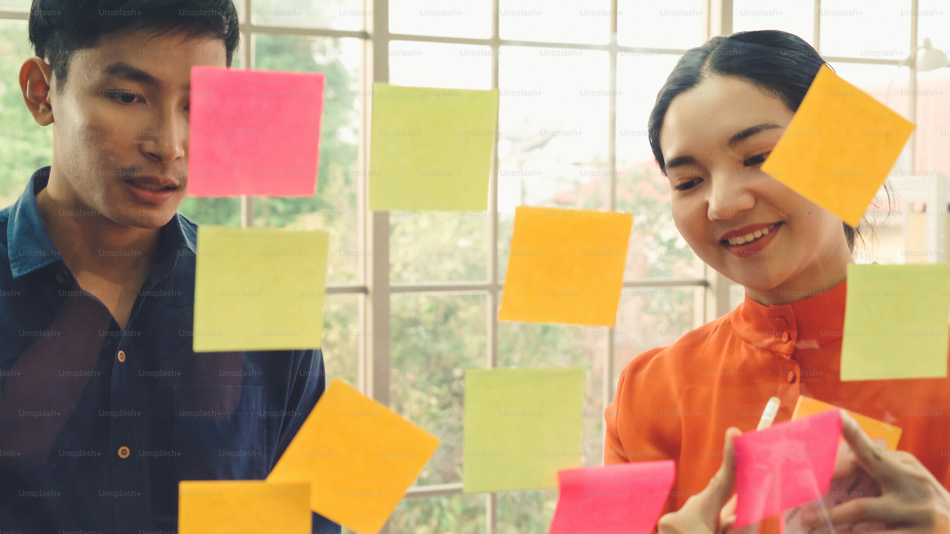 La gente de negocios trabaja en el tablero de planificación de proyectos en la oficina y tiene una conversación con un amigo compañero de trabajo para analizar el desarrollo del proyecto. Usan notas adhesivas colocadas en una pared de vidrio para organizarlo.