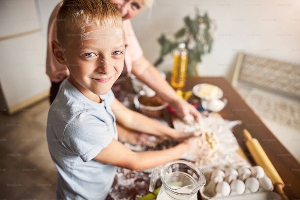 Selective focus photo of cute smiley boy making dough with his grandma and looking at the camera