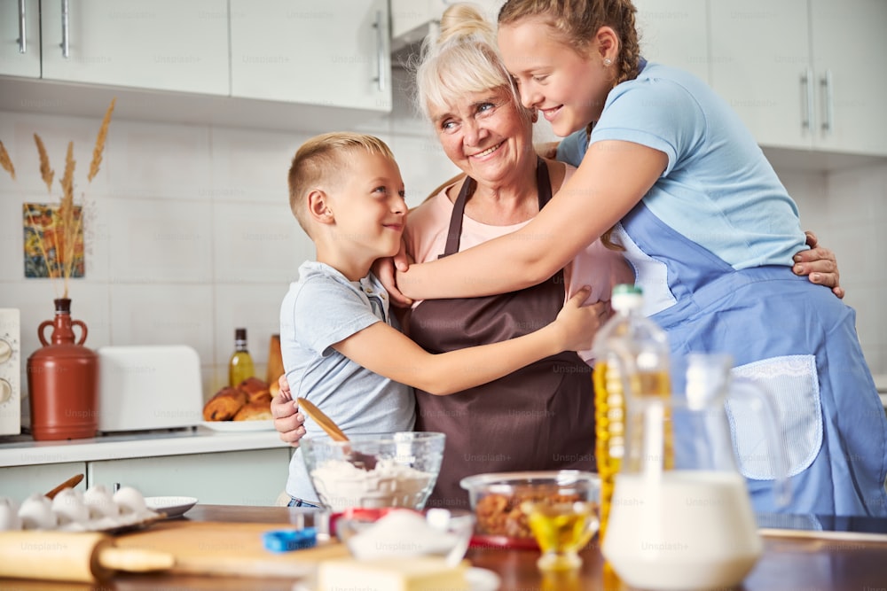 Abuela feliz sonriendo y abrazando a sus dos nietos mientras está de pie en una acogedora cocina