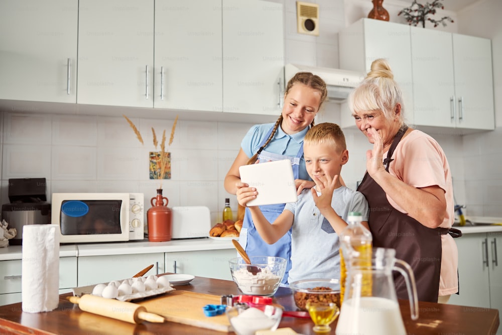 Joyful senior lady and two grandchildren smiling and waving at tablet screen while standing in the kitchen