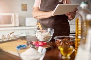 Foto ritagliata di donna non rivestita in un grembiule in piedi vicino al tavolo della cucina mentre tiene in mano una tavoletta