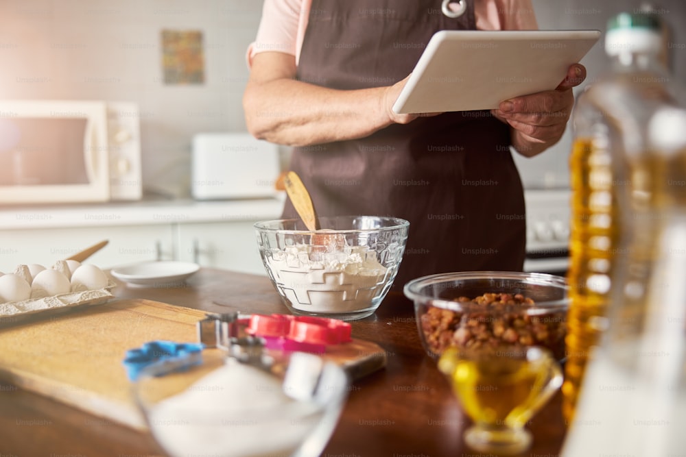 Cropped photo of unreconised woman in an apron standing near kitchen table while holding a tablet