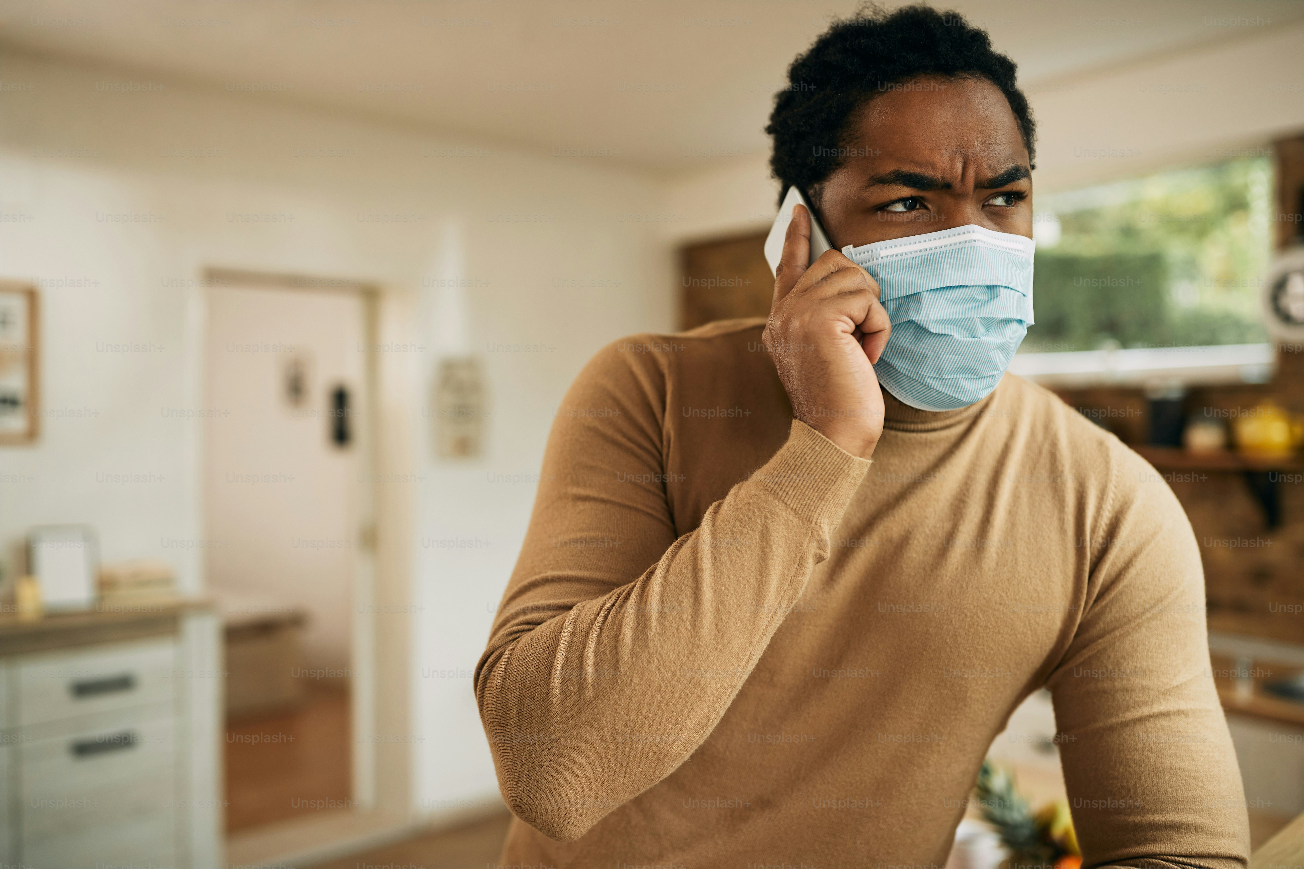 African American man with face mask communicating over mobile phone from home during COVID-19 pandemic.