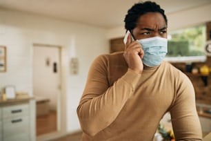 African American man with face mask communicating over mobile phone from home during COVID-19 pandemic.