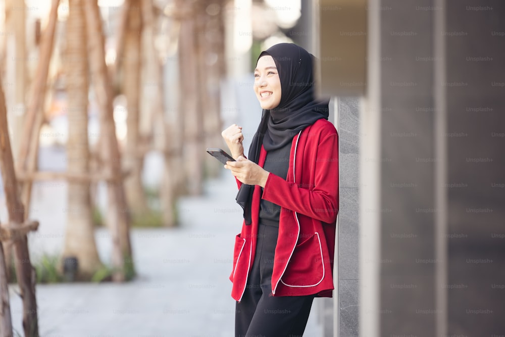 Jovem muçulmana que celebra o sucesso segurando o telefone celular em pé na rua