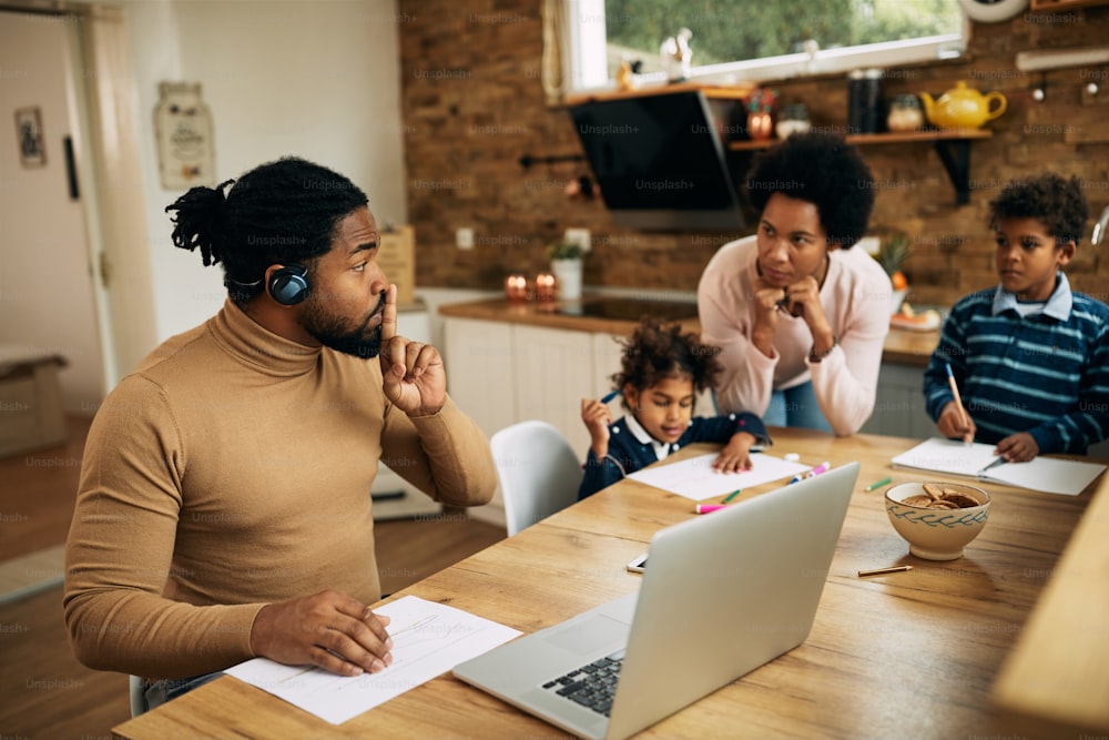 아프리카계 미국인 남성이 노트북으로 화상 통화를 하고 집에서 일하는 동안 아내와 아이들을 침묵시키고 있습니다.