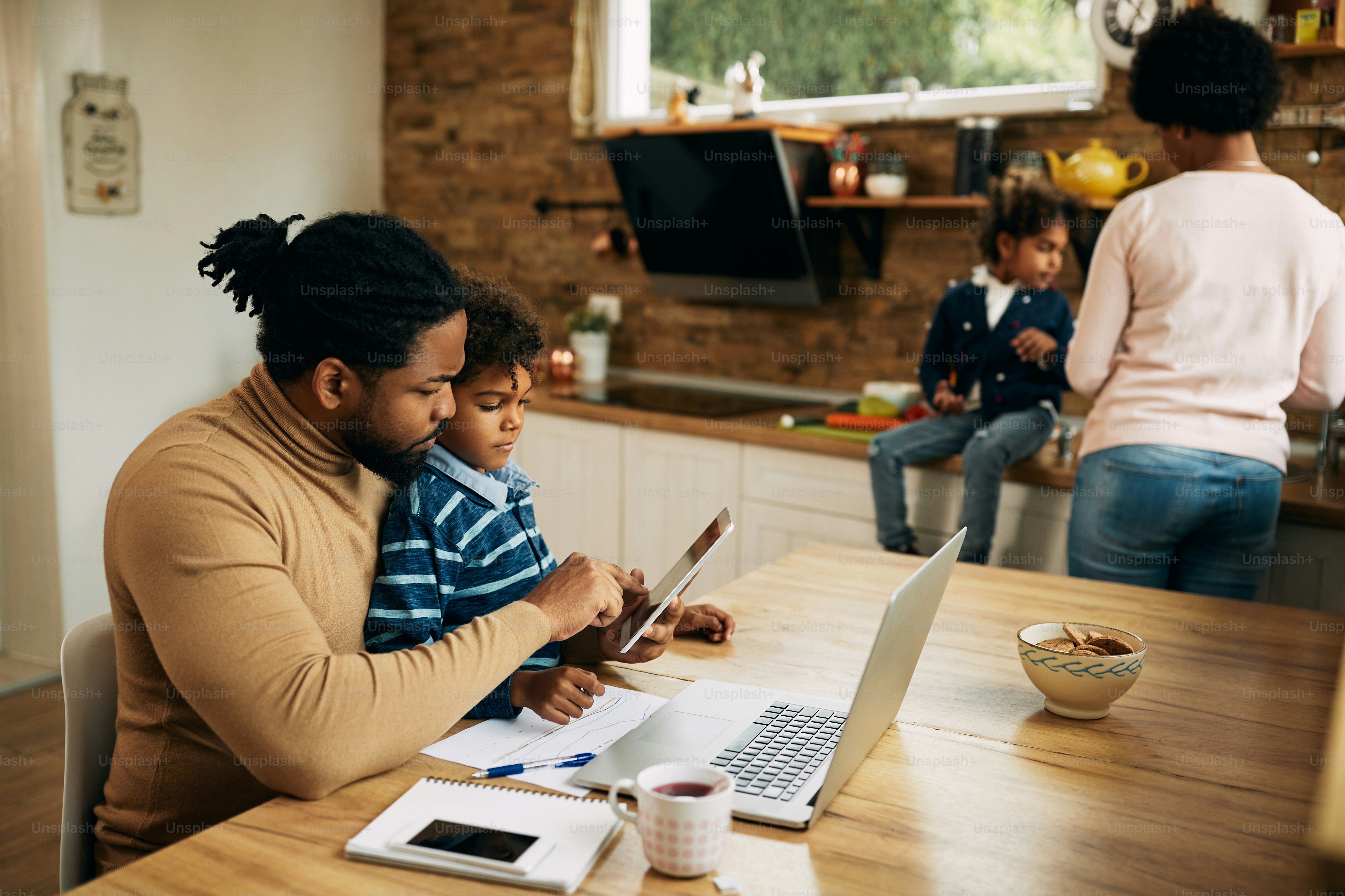 Black working father using digital tablet while son is sitting on his lap. Mother and daughter are in the background.