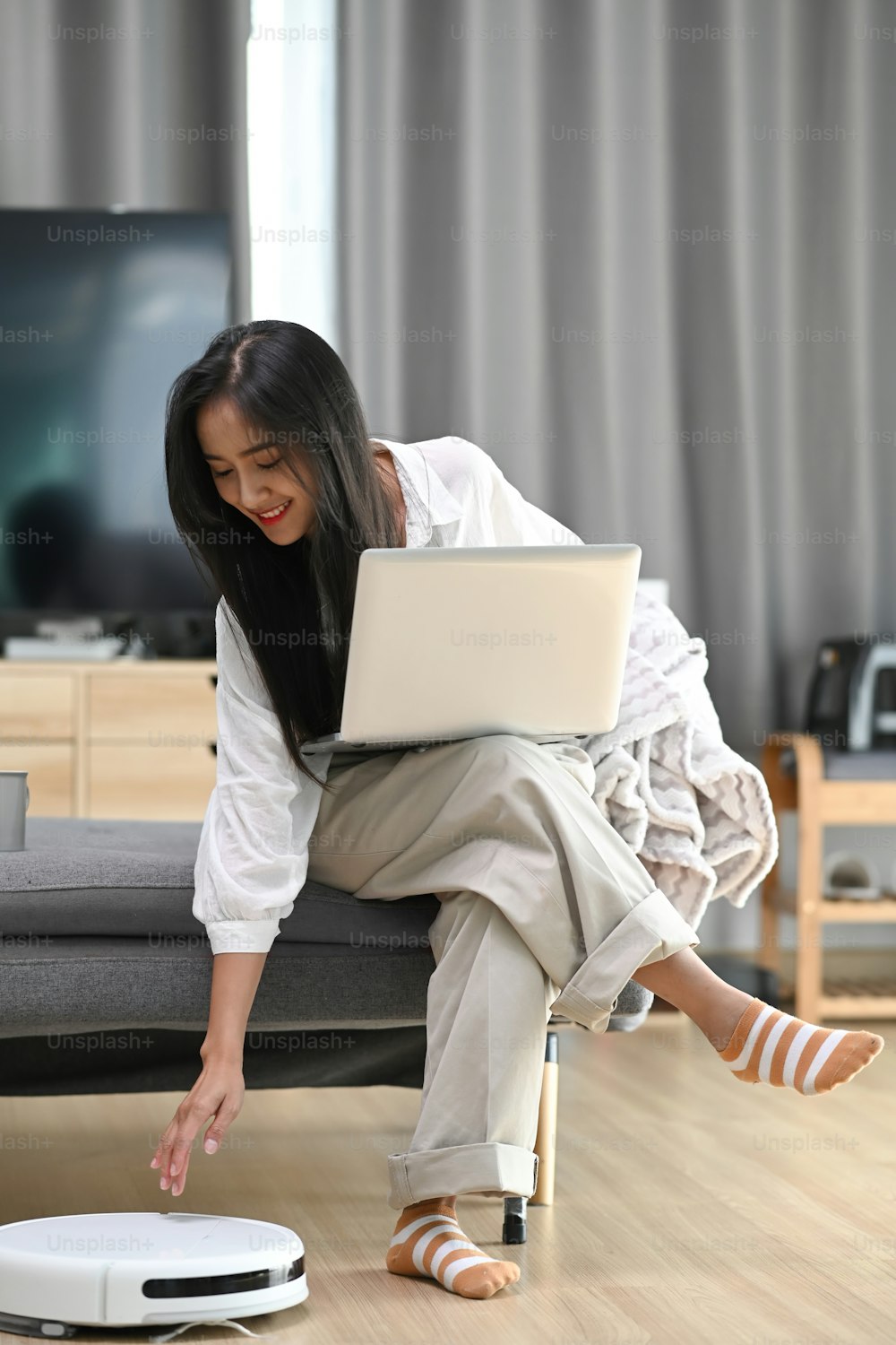 Mujer feliz con computadora portátil sentada en el sofá y robot aspirador limpiando la habitación en casa.