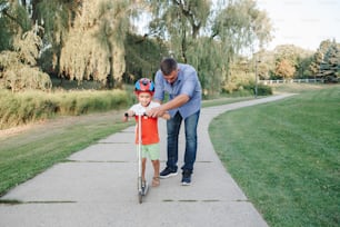 Caucasian father dad training teaching boy son to ride scooter. Preschooler child kid in helmet with bike on backyard park road. Seasonal summer child family activity outdoor.