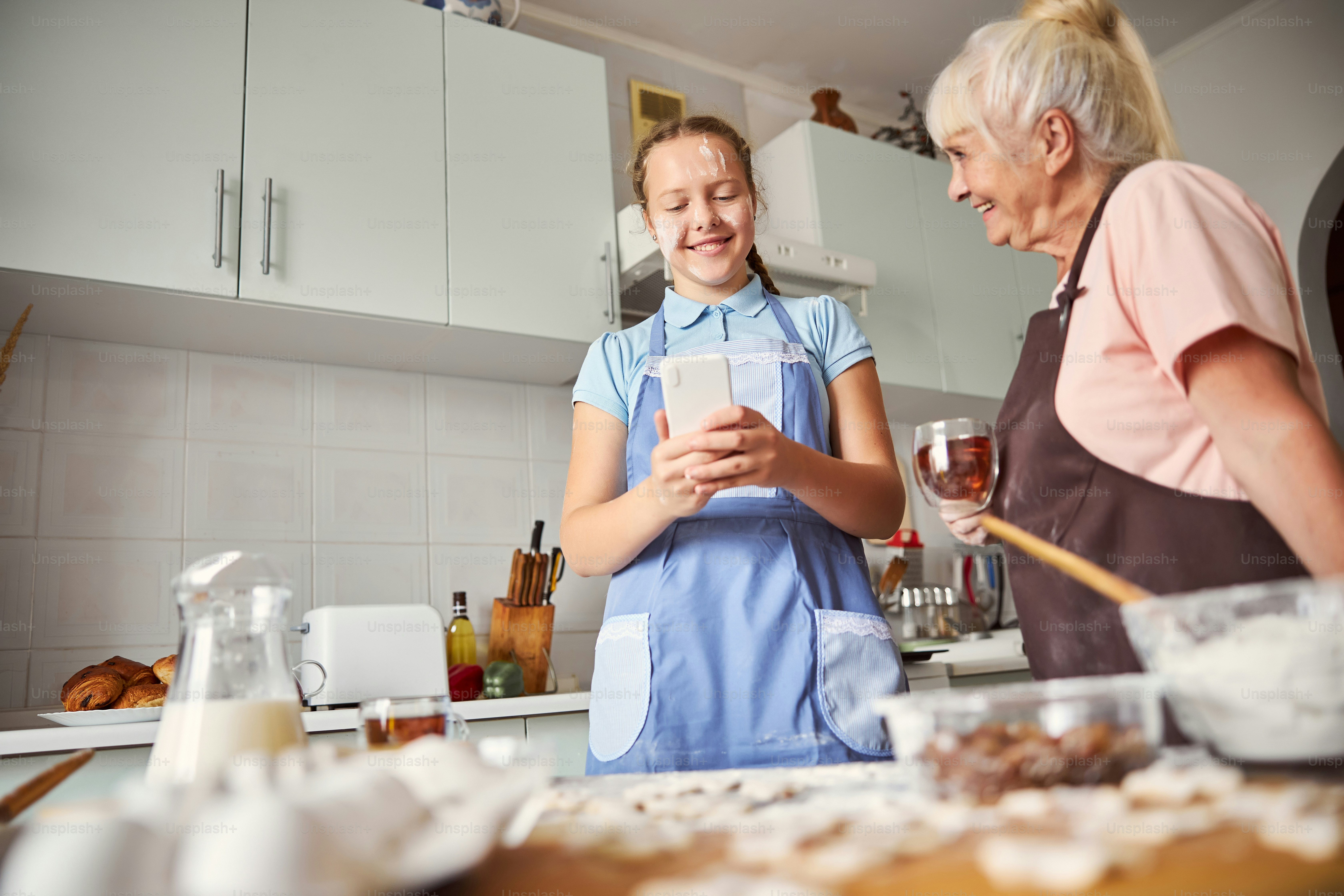 Joyous girl with braids looking at her smartphone while her grandma watchine her