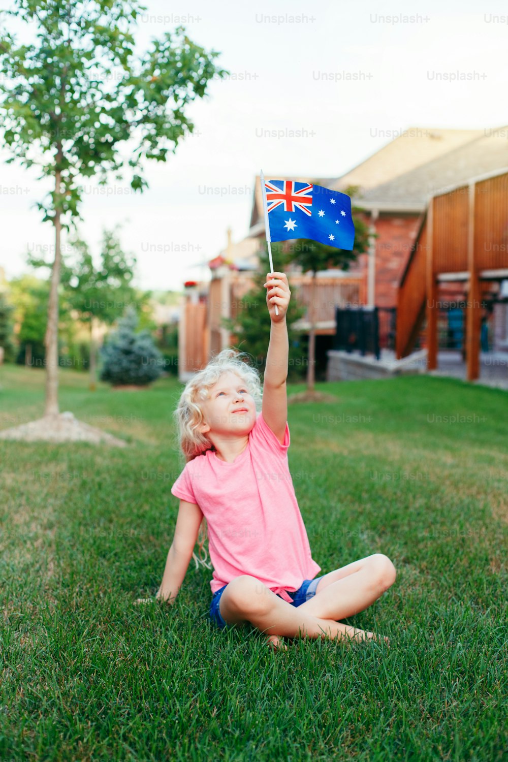 Feliz chica rubia caucásica sosteniendo la bandera australiana. Niño sonriente sentado en la hierba en el parque ondeando la bandera de Australia. Niño ciudadano que celebra el feriado del Día de Australia en enero al aire libre.
