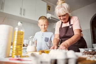 Enthusiastic aged lady being concentrated on cooking while her grandson watching her