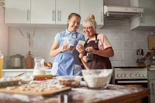 Joyous girl and happy eldelry lady looking at smartphone while having tea in cozy kitchen