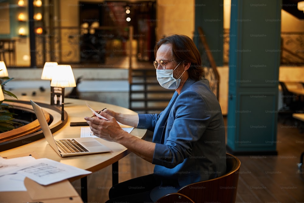 Responsible brunette man in smart blazer wearing a mask while working remotely from a comfortable lounge