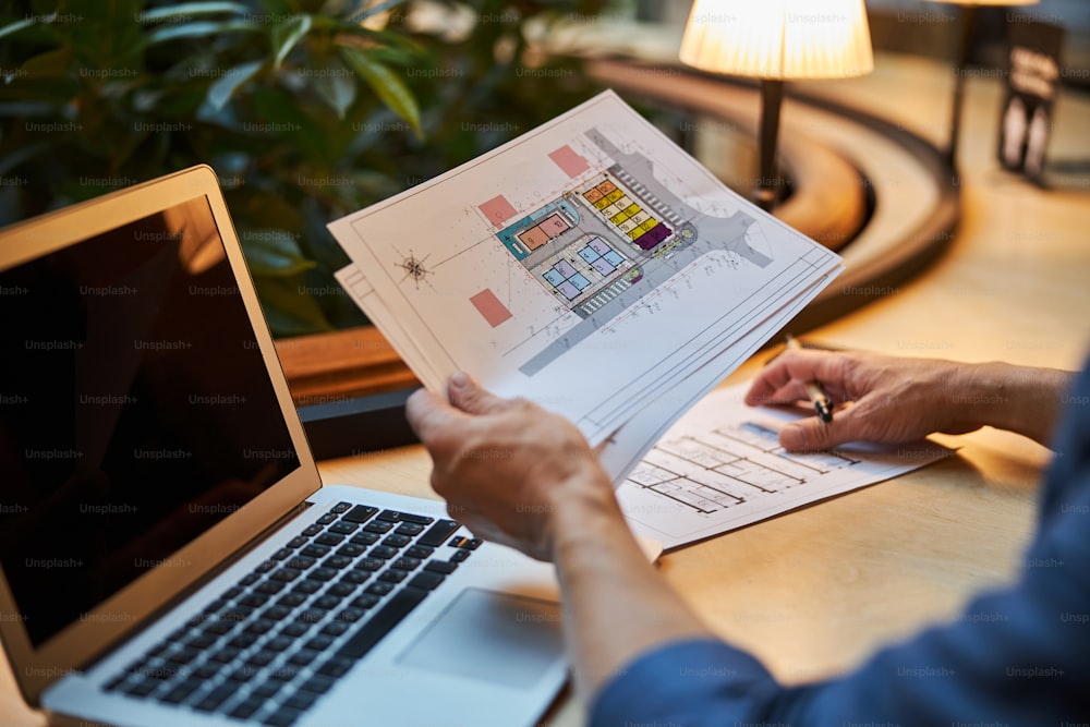 Cropped photo of man sitting at desk in front of laptop and holding some building plan prints