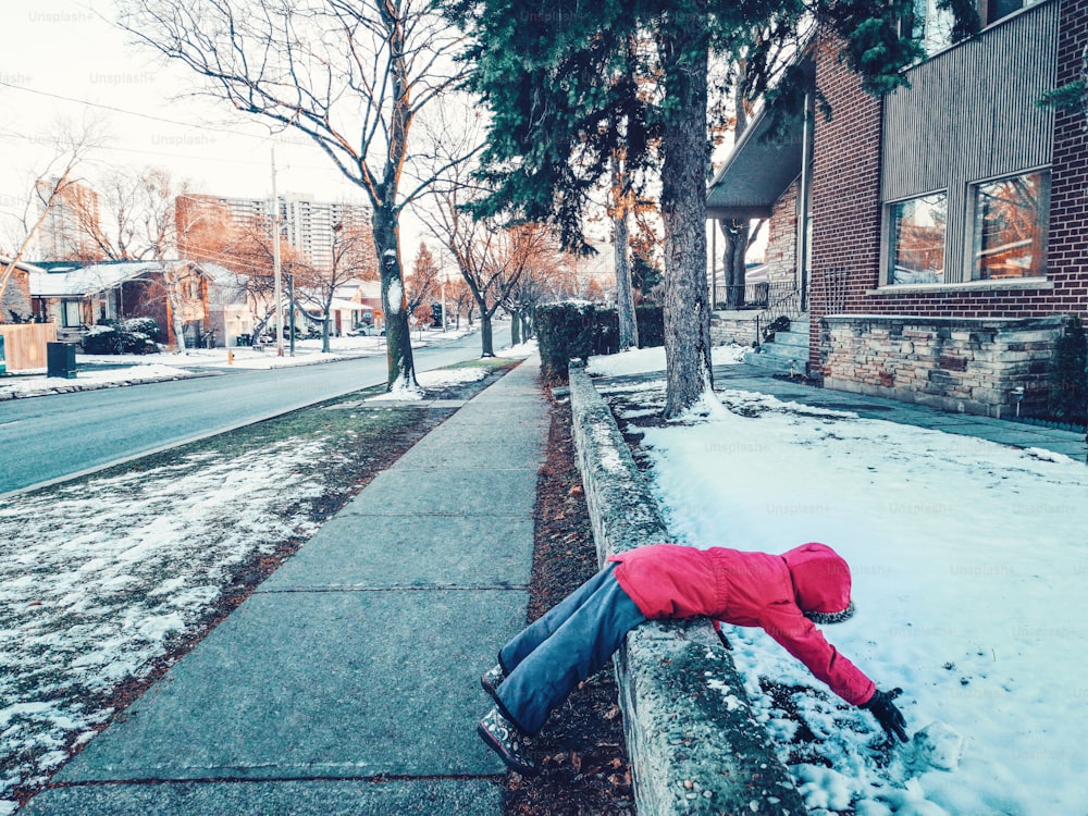 Funny child girl in warm clothes jacket lying in snow on city street. Kid having fun making snowball on cold winter day. Naughty cranky kid playing outside. Hilarious funny weird kid outdoor.