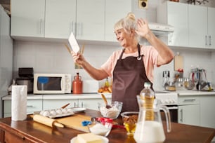 Excited senior woman smiling and waving at tablet screen while standing in her well-furnished kitchen