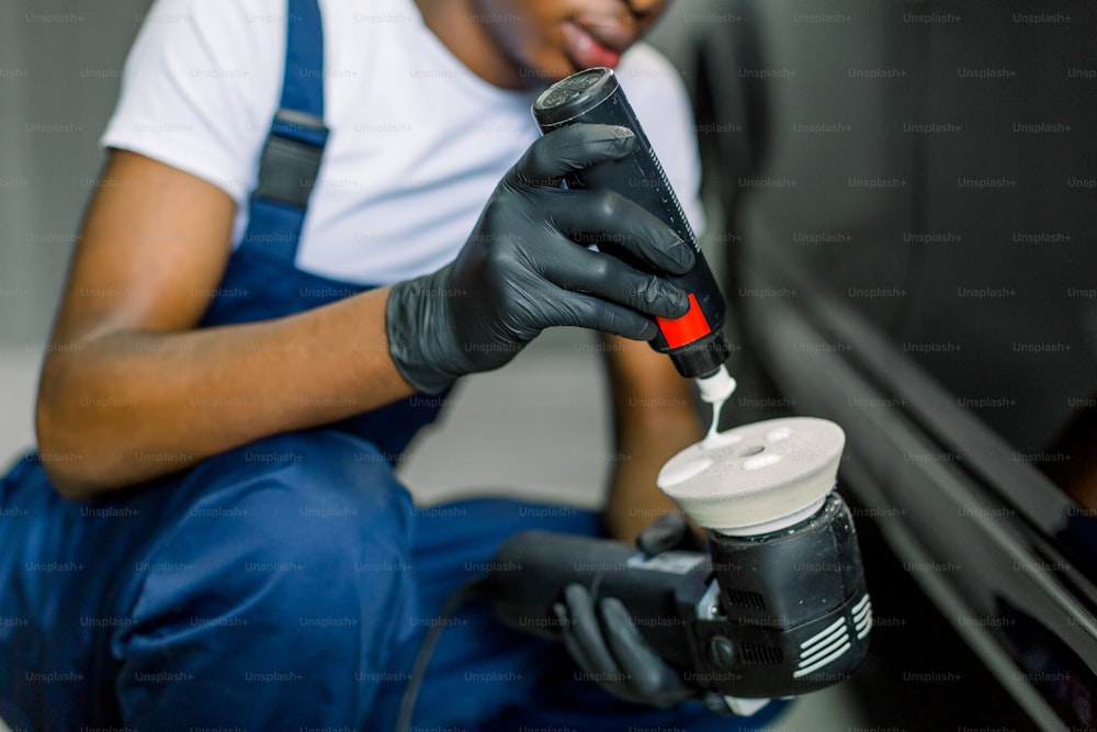 Cropped close up image of hands of African male auto service worker, pouring special polish wax or cream on the orbital polish machine to polish the black car on the background.
