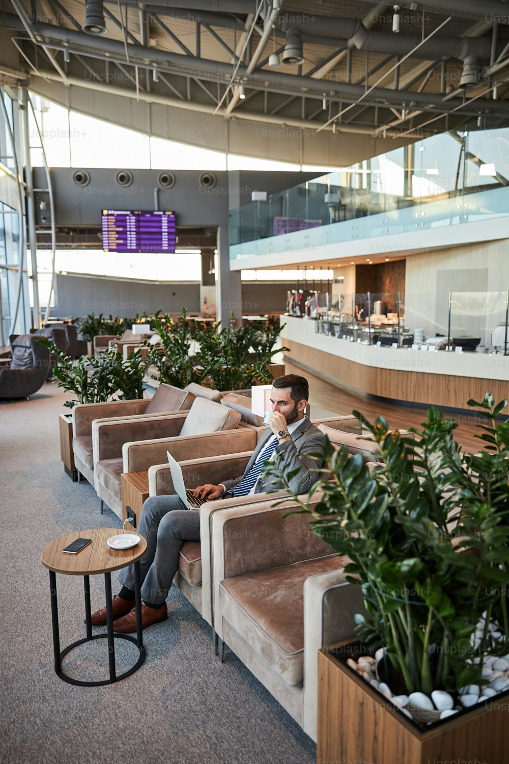 Homem bonito calmo em um terno sentado com um laptop em um salão de negócios e tomando um gole de seu copo