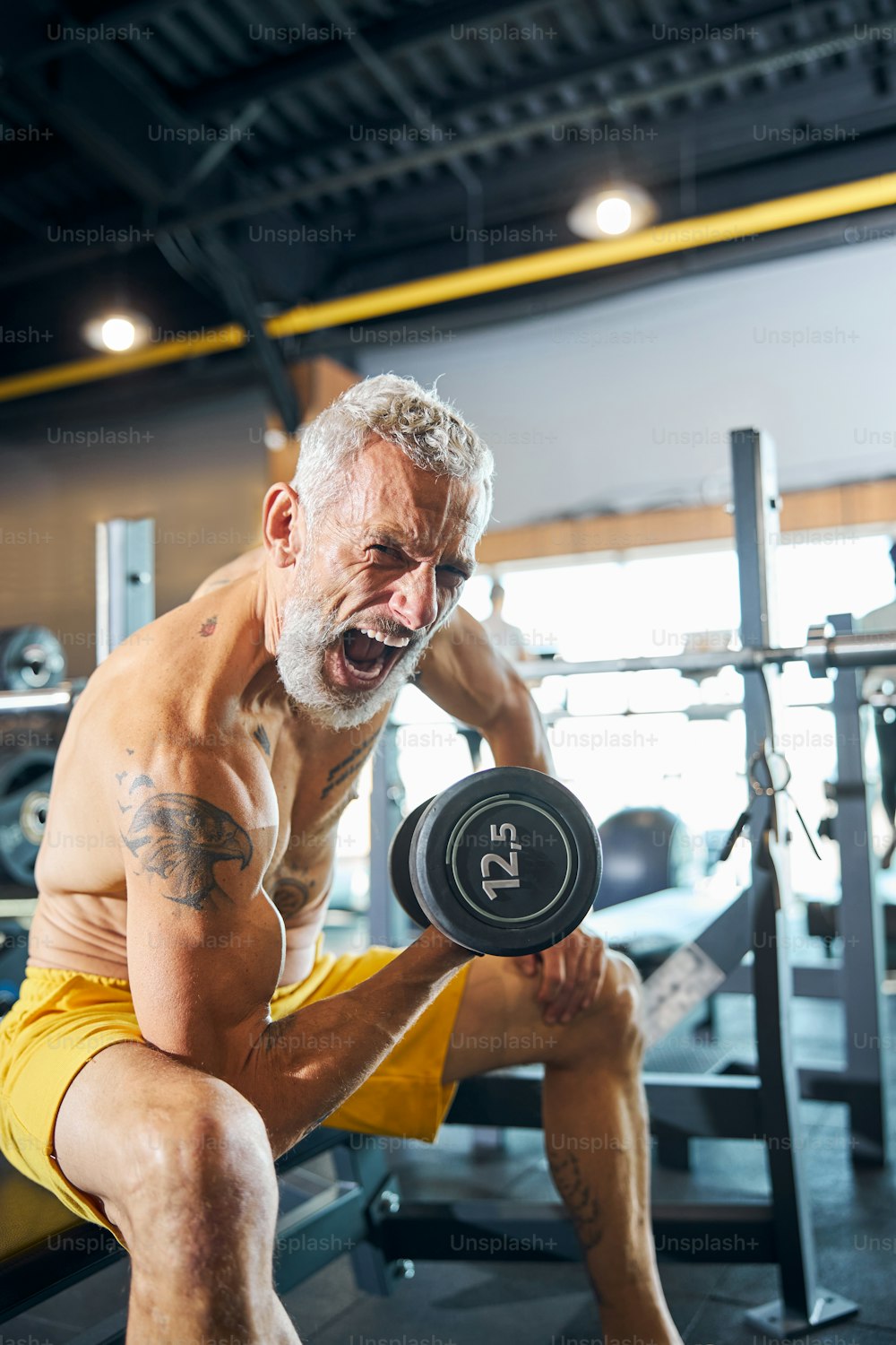 Gray-headed shirtless professional Caucasian male athlete screaming during the strength workout in a fitness center