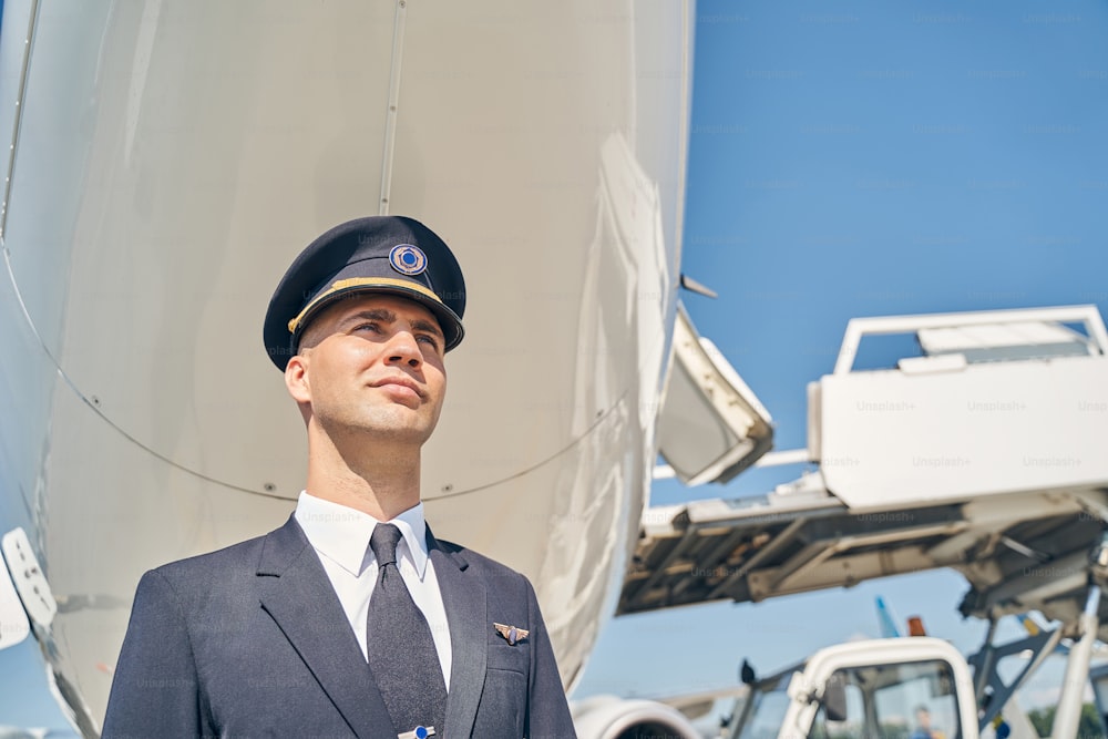 Portrait d’un jeune pilote sérieux debout seul sous le fuselage de l’avion à l’aérodrome