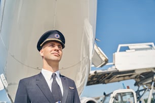 Ritratto di un giovane volatore serio in piedi da solo sotto la fusoliera dell'aereo all'aerodromo