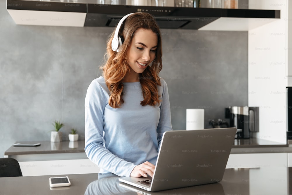 Portrait of a joyful young woman listening to music with headphones and using laptop computer while standing at the kitchen table at home