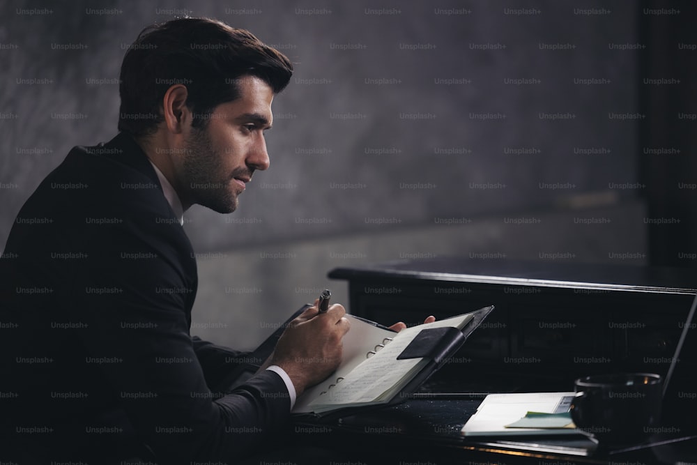 Businessman writes in a notebook while sitting at a desk, dark tone