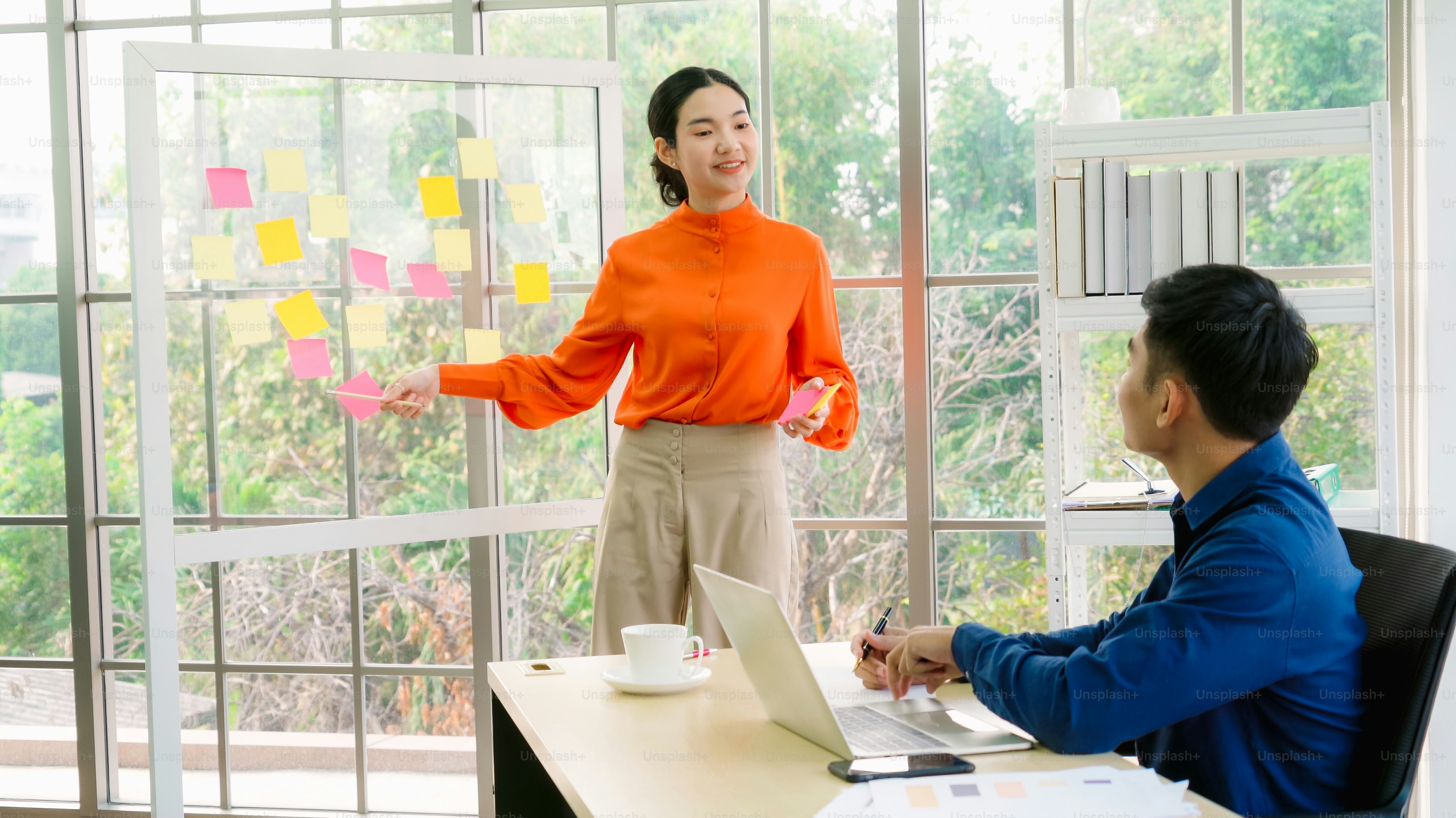 Les gens d’affaires travaillent sur le tableau de planification de projet au bureau et ont une conversation avec un ami collègue pour analyser le développement du projet. Ils utilisent des notes autocollantes affichées sur le mur de verre pour l’organiser.