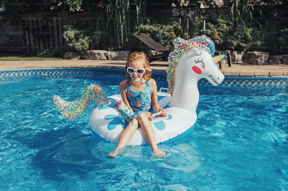 Süßes entzückendes Mädchen in Sonnenbrille mit Getränk auf aufblasbarem Ring Einhorn liegend. Kind genießt Spaß im Schwimmbad. Sommer Wasseraktivität im Freien für Kinder.
