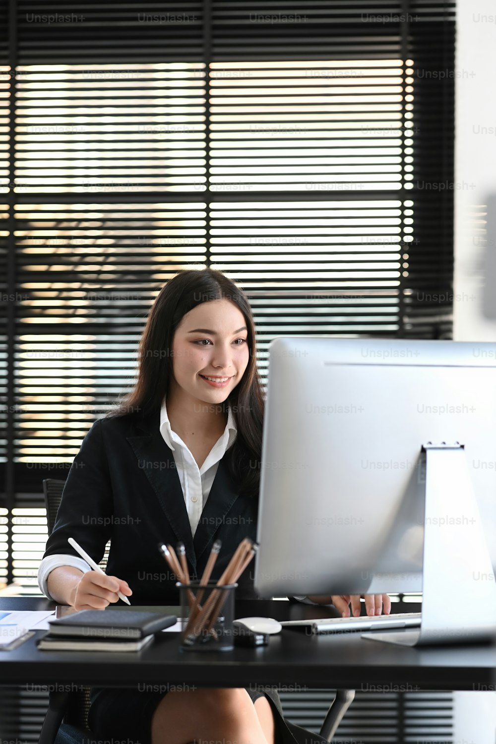 Portrait of female office worker in black suit working on computer and sitting in modern office.