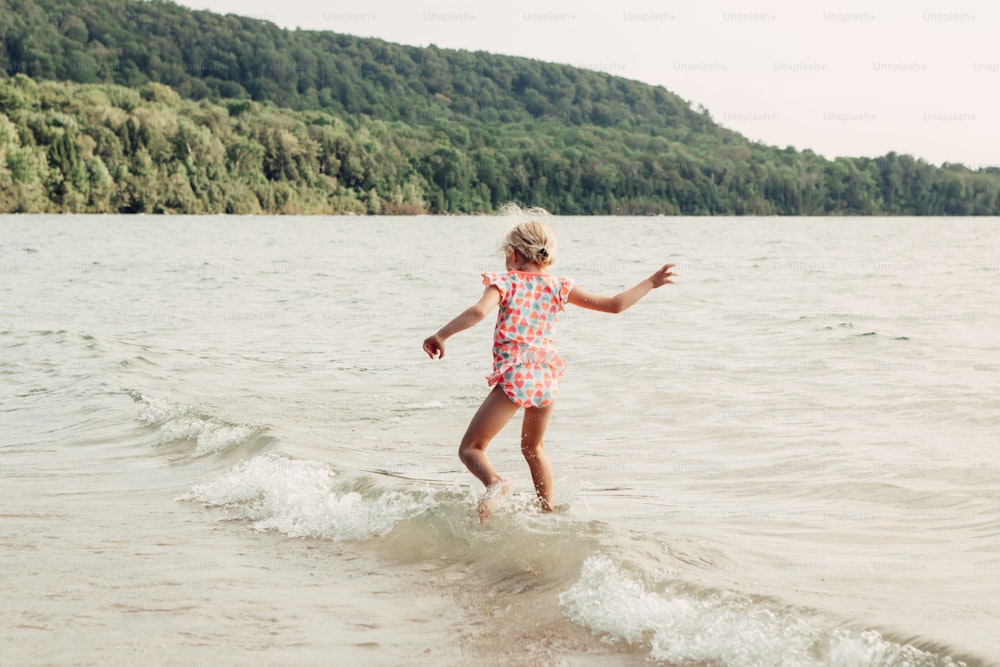 Menina caucasiana engraçada engraçada que caminha nas ondas do mar do lago ao pôr do sol. Criança pulando na água na praia. Autêntico estilo de vida real infância feliz. Diversão de verão atividade sazonal ao ar livre. Vista de trás.