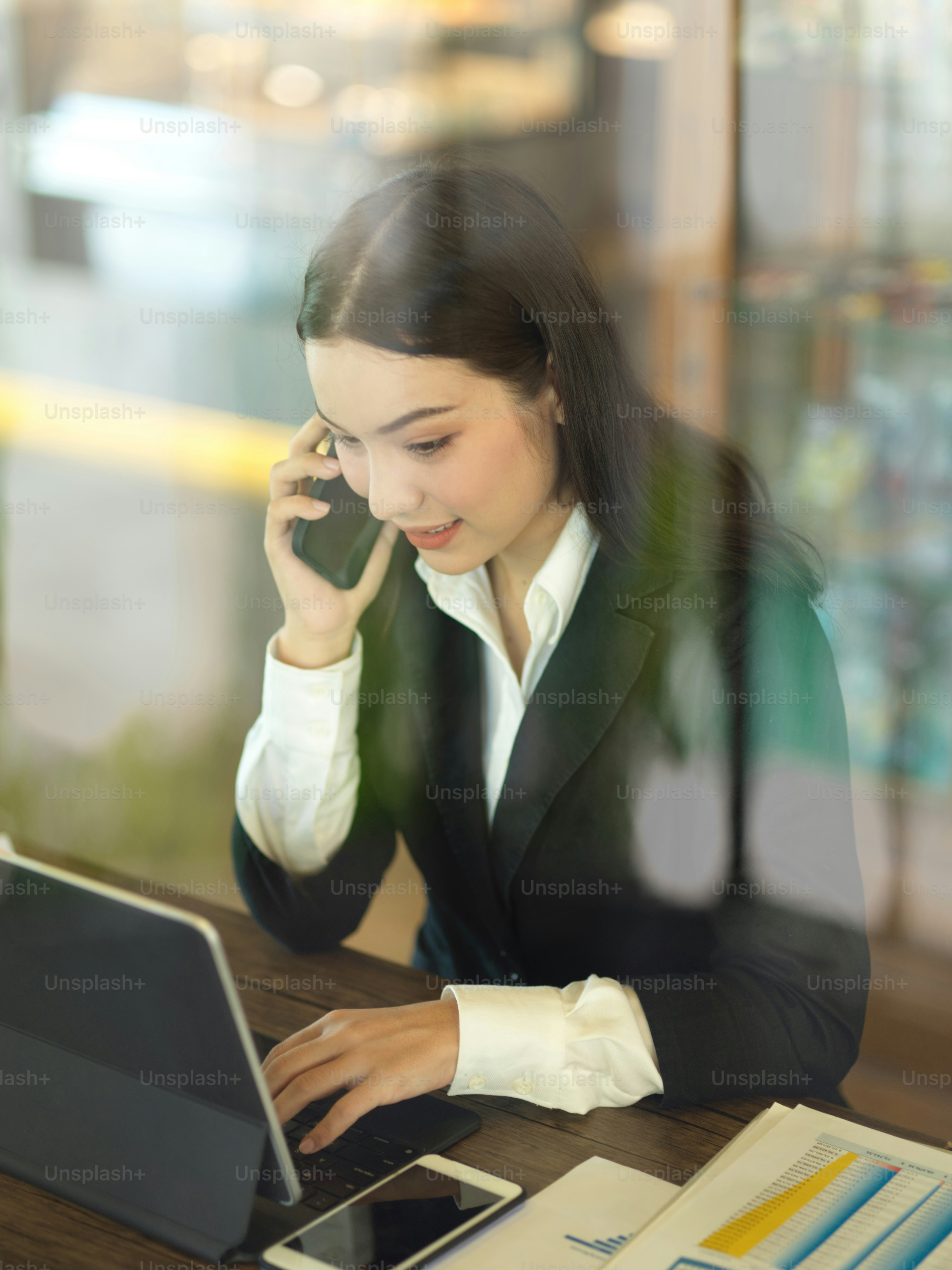 View through glass wall of businesswoman talking on the phone and working with tablet in office