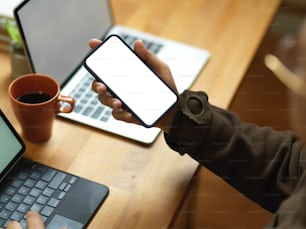 Cropped shot of male hand holding smartphone while working with laptop and tablet in office room