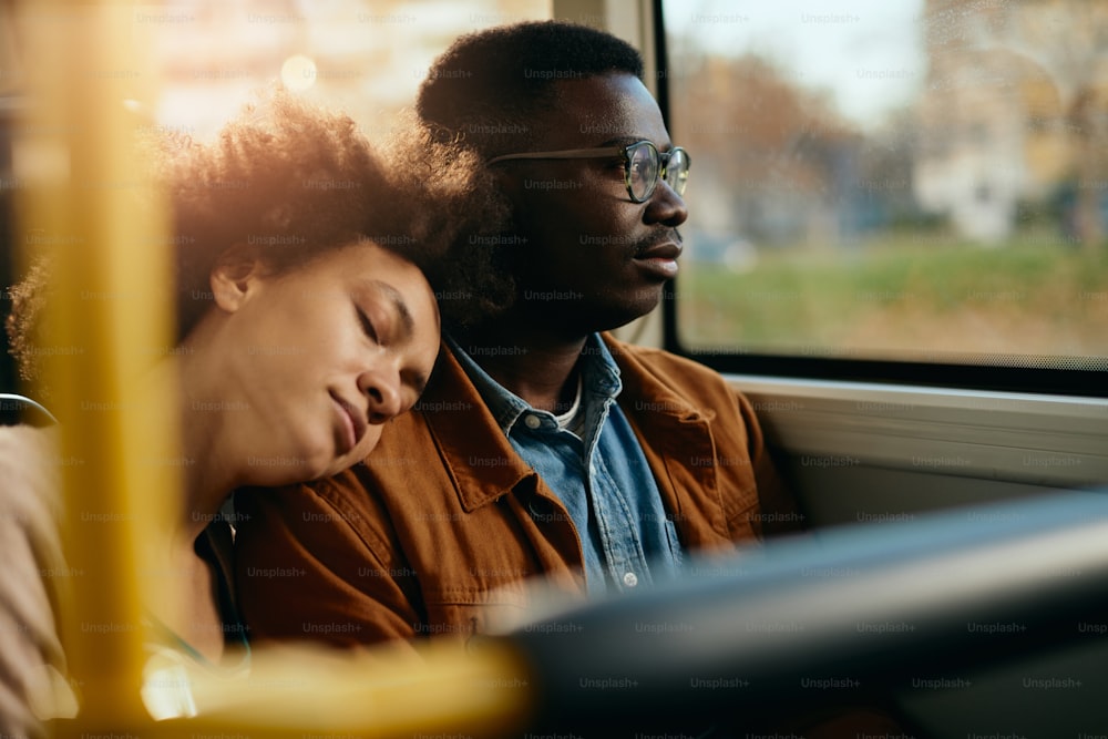 Uomo nero pensieroso che guarda attraverso il finestrino mentre la sua ragazza si addormenta sulla sua spalla in un autobus.