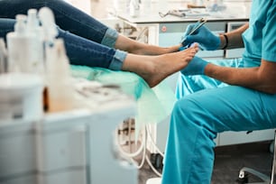 Cropped head portrait of unrecognized patient sitting on the medical chair while doctor in blue cloves examining feet in beauty center