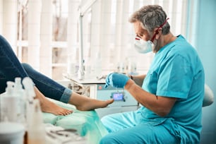 Side view portrait of self confident European male in uniform and gloves removing pain in toes on female feet in medical center