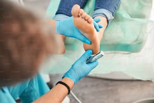 Close up portrait of pedology beautician in blue gloves cleaning skin of client leg from callus and corn with professional tool.