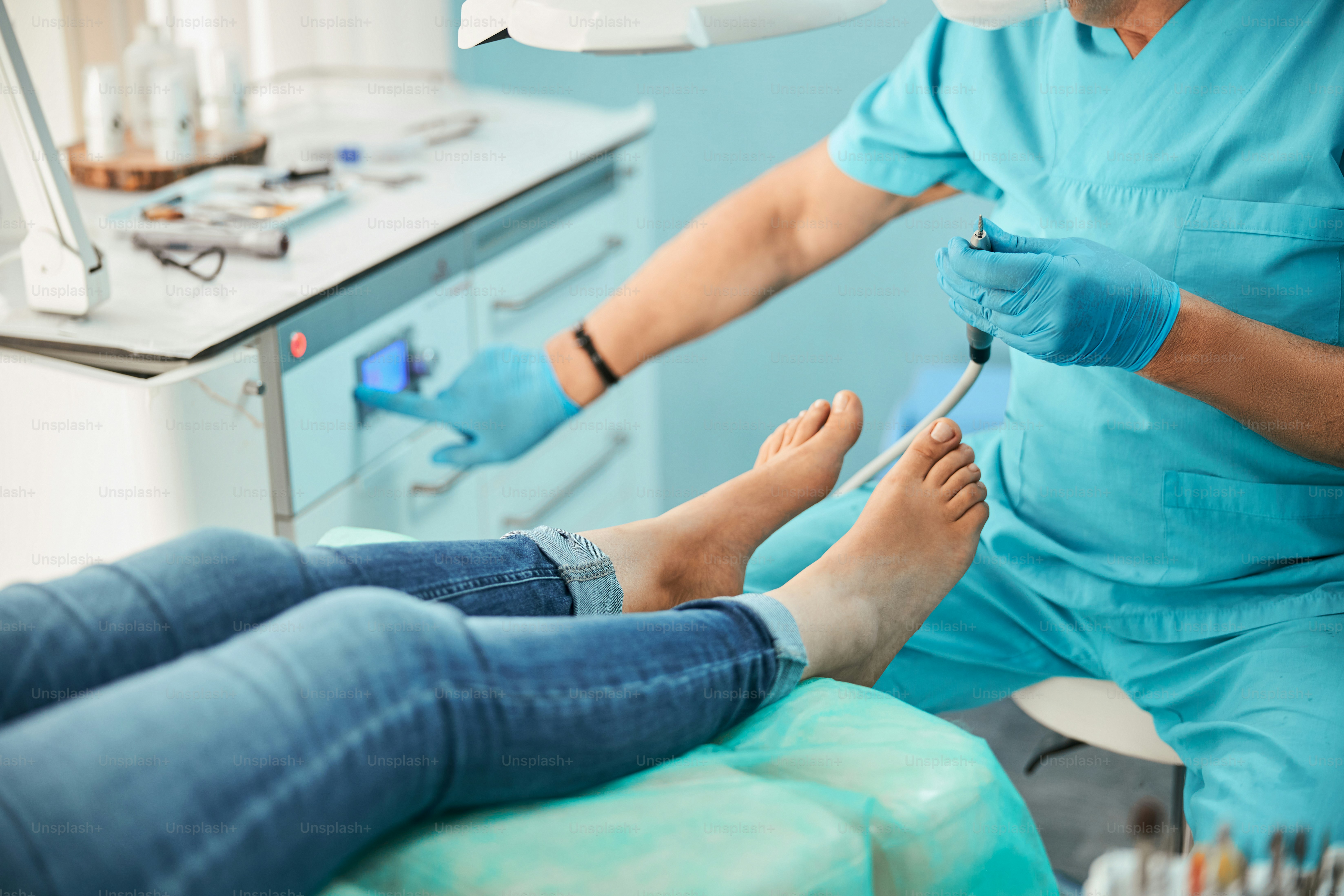 Female legs on medical chair while pedicurist in blue uniform and gloves doing professional medical pedicure procedure in beauty salon with nail electrical tools
