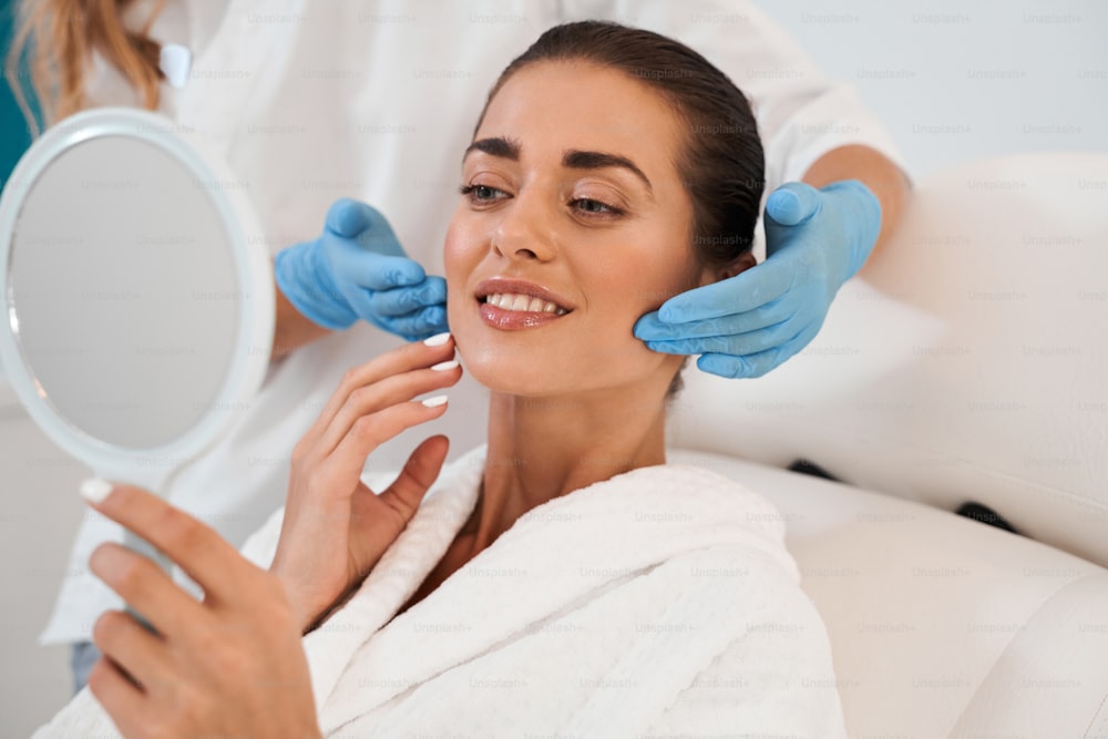 Young woman at beauty clinic cosmetology service sitting on medical chair while female doctor wearing gloves holding her face examining skin with mirror concentrated close-up