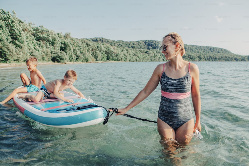 Caucasian woman parent riding kids children boys on paddle sup surfboard in water. Modern outdoor summer fun family activity. Individual aquatic recreation sport hobby. Healthy lifestyle.