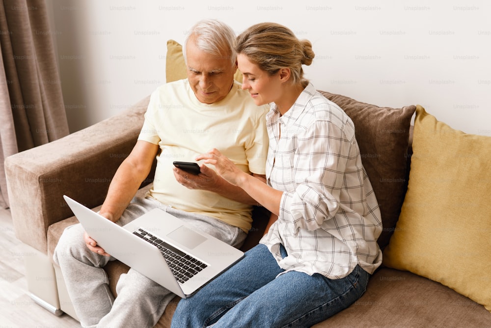 Young woman explaining to elderly man how to use laptop and smatphone