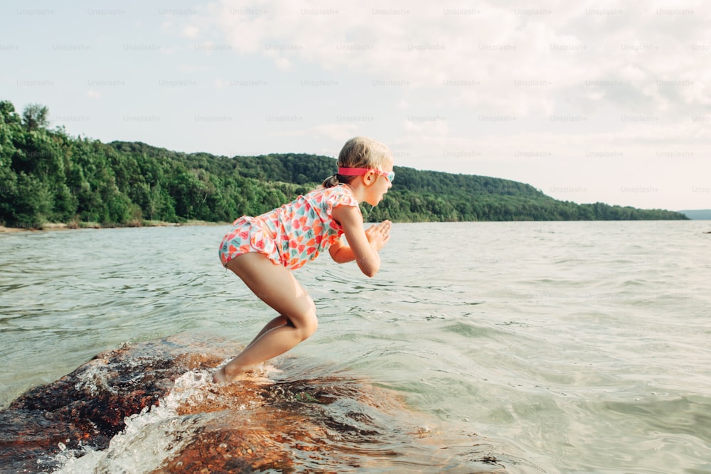 Cute funny Caucasian girl diving in lake with underwater goggles. Child swimming in water on beach. Authentic real lifestyle happy childhood. Summer fun outdoor aquatic seasonal sport activity.