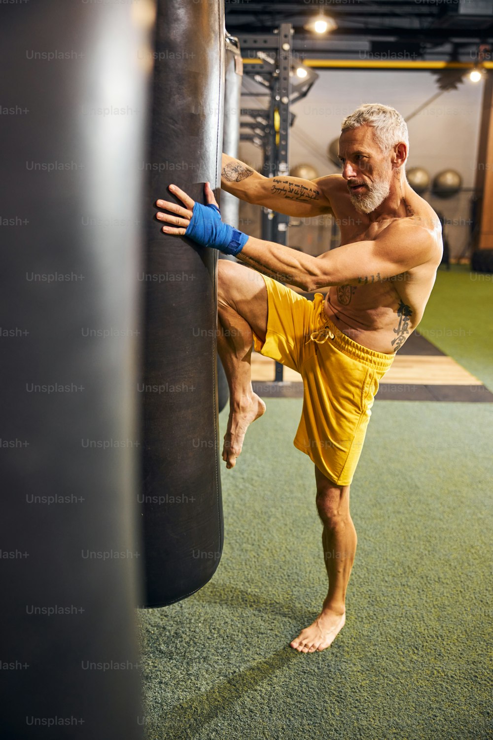 Full-sized portrait of a gray-headed bearded barefoot shirtless Caucasian male athlete kneeing the punching bag
