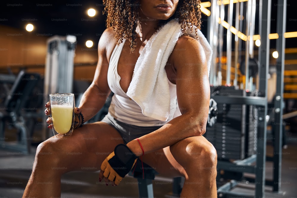 Foto recortada de una deportista con un vaso de batido de proteínas sentada en el gimnasio