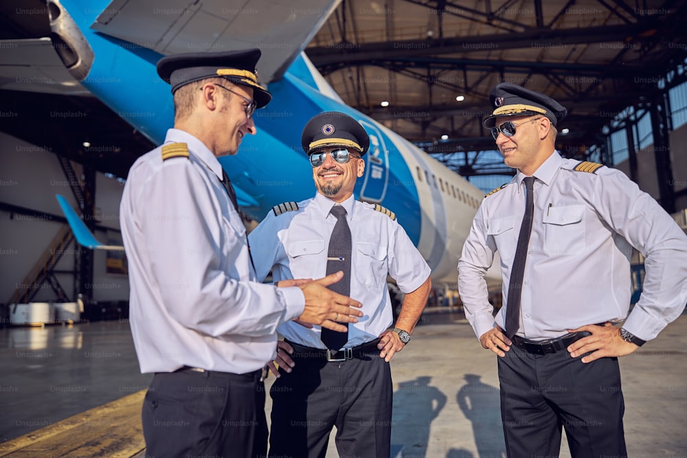 Retrato de cerca de pilotos guapos sonrientes en traje de negocios y gafas de sol hablando juntos mientras pasan tiempo en el hangar de aviación cerca del avión de pasajeros