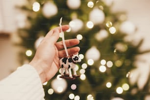 Bonne année 2021 ! Main tenant un jouet de vache ou de taureau sur fond de belles lumières d’arbre de Noël bokeh, espace pour le texte. Décor de figurine de vache mignonne sur sapin de Noël, symbole du nouvel an 2021