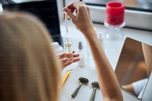 Cropped head back view portrait of young female holding plastic container with serum or essential oil in hand above the white desk