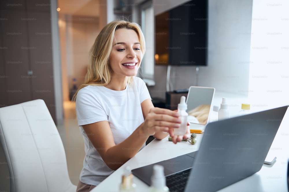 Waist up portrait of smiling blonde Caucasian woman sitting at the laptop and advertising skin cosmetic on the video call in home apartment