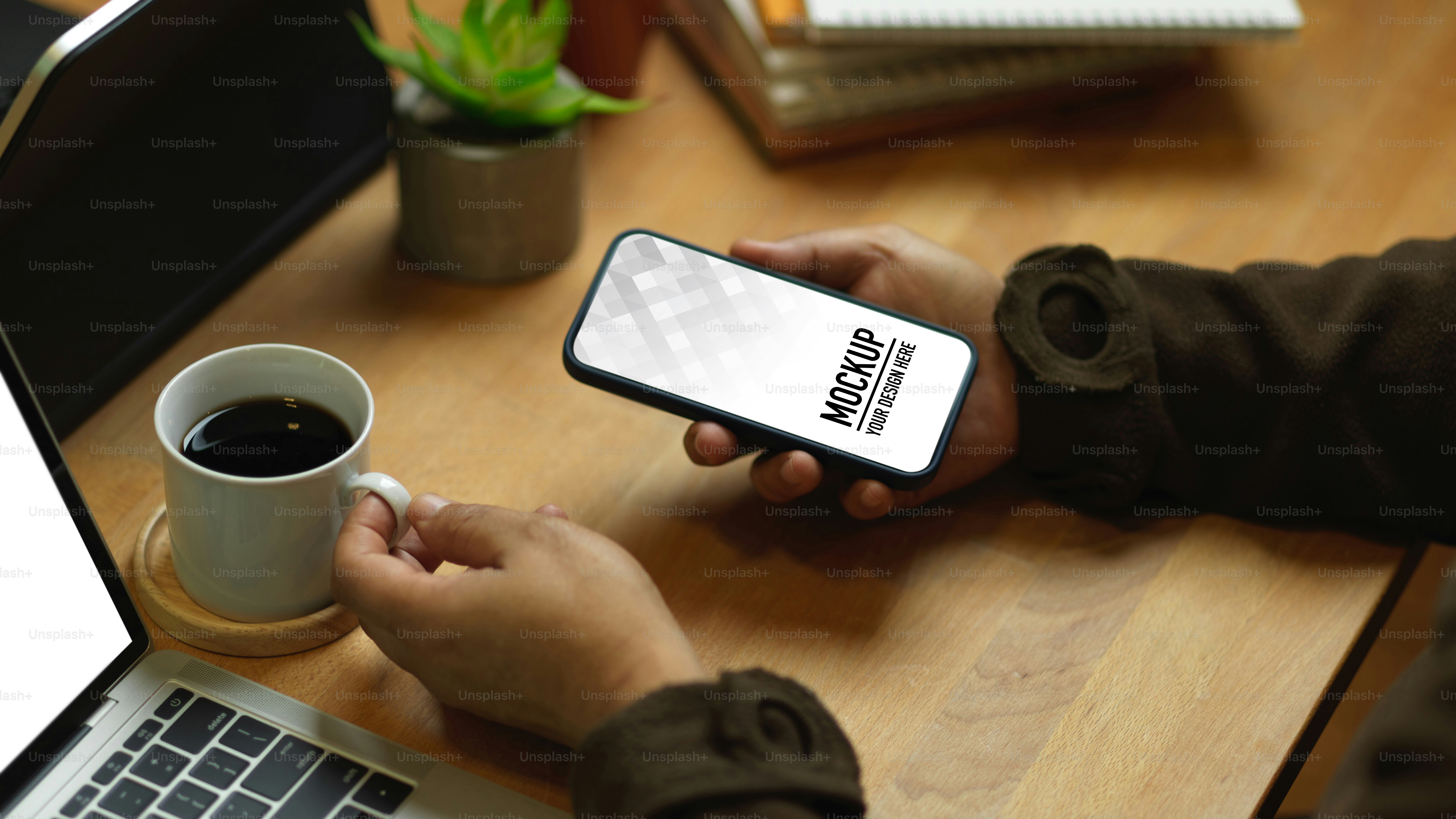 Colpo ritagliato di mano maschile che tiene lo smartphone e la tazza di caffè sul tavolo da lavoro in legno, percorso di ritaglio